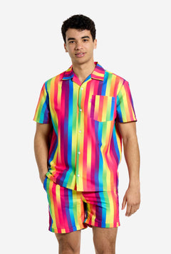 Een man draagt een verticaal gestreepte regenboogkleurige zomerset, bestaande uit een overhemd en een korte broek. Hij lacht in de camera en houdt één hand in zijn zak.