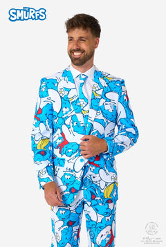 Lachende man gekleed in een kleurrijk herenpak van de Smurfen met een all-over cartoonprint in de kleuren blauw, wit, rood en geel, inclusief bijpassende stropdas en wit overhemd, staande tegen een effen achtergrond.