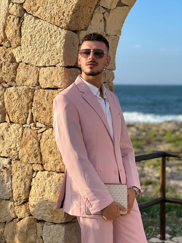 Man in een roze pak staande tegen een stenen muur met uitzicht op de oceaan