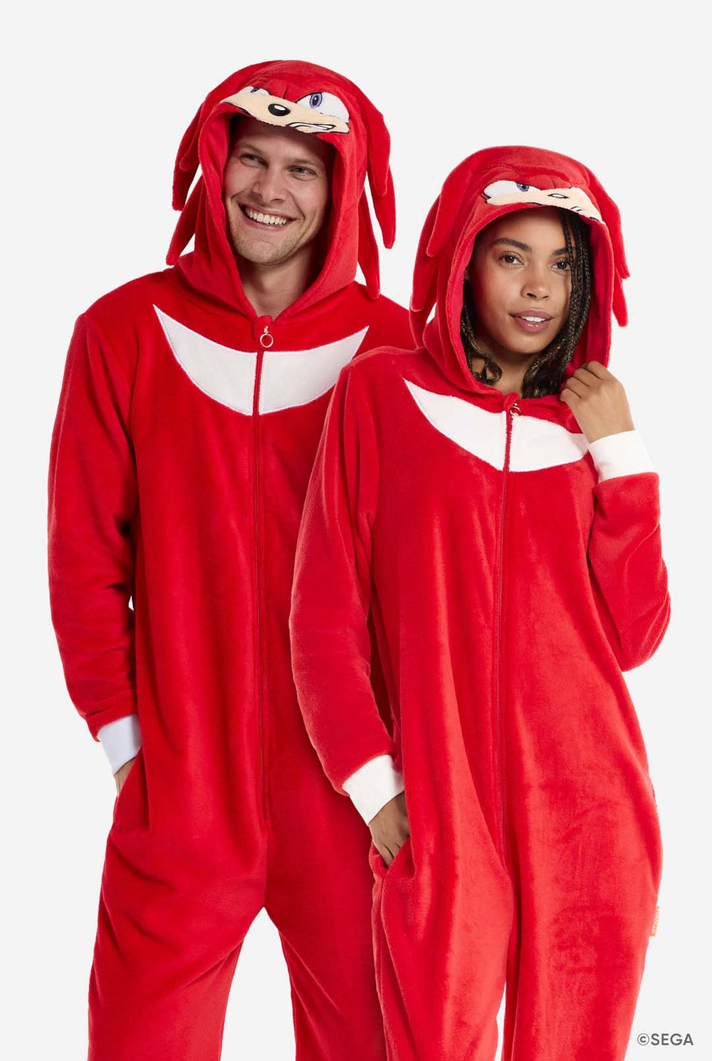 Een man en een vrouw dragen een felrode Knuckles the Echidna onesie voor volwassenen van OppoSuits. De leuke en comfortabele onesie heeft een ritssluiting aan de voorkant en een capuchon die is ontworpen om Knuckles' gezicht te imiteren, compleet met expressieve ogen en flodderige dreadlocks. Het pak is overwegend rood, met een wit halvemaanpatroon op de borst, een verwijzing naar Knuckles' iconische look uit de Sonic the Hedgehog-videogameserie.