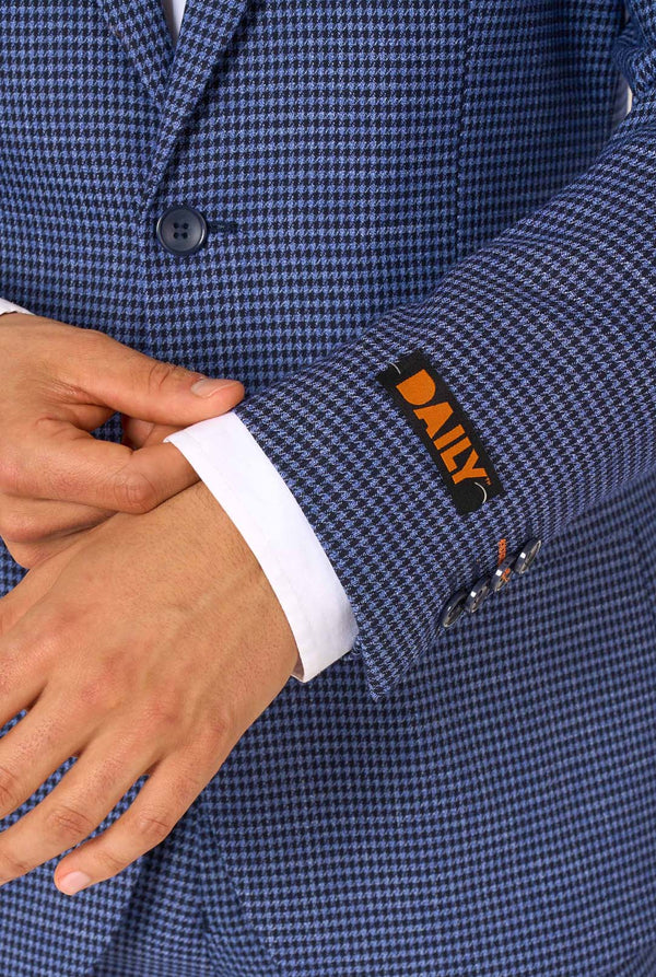 Een man draagt een marineblauw en blauw geruit pak met een wit overhemd. Het pak heeft een moderne pasvorm met twee knopen en klepzakken. Hij staat tegen een effen witte achtergrond. Close up van de mouwen