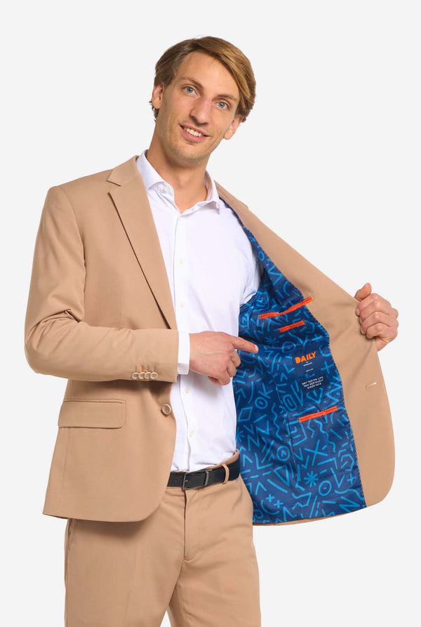 Afbeelding met lichtgrijze achtergrond en man die donker zand mannenpak en wit overhemd draagt. Pak bestaat uit blazer en broek. Houd een zijde van het jasje open.