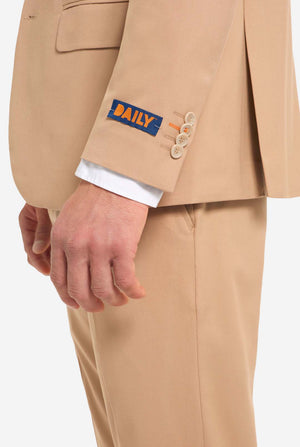 Afbeelding met lichtgrijze achtergrond en man die donker zand mannenpak en wit overhemd draagt. Pak bestaat uit blazer en broek. Zicht vanaf de zijkant.