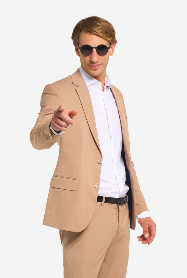 Afbeelding met lichtgrijze achtergrond en man met zonnebril, donker zandkleurig mannenpak en wit overhemd. Pak bestaat uit blazer en broek. Wijzend naar de camera.