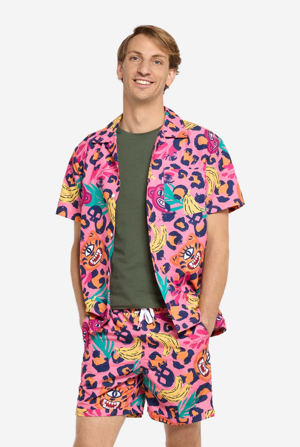Een man die een levendige tweedelige zomeroutfit draagt ​​met een opvallende tropische print. Het overhemd met korte mouwen en de bijpassende shorts hebben een levendig ontwerp met neonroze, oranje en gele luipaardprints, bananen, groene palmbladeren en eigenzinnige cartoongezichten. De outfit is nonchalant gestyled met een groen T-shirt eronder en is perfect voor zomerfeestjes, vakanties of plezier bij het zwembad.