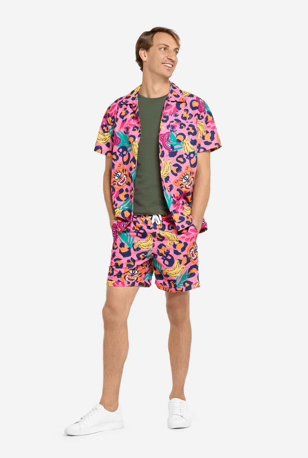 Een man die een levendige tweedelige zomeroutfit draagt ​​met een opvallende tropische print. Het overhemd met korte mouwen en de bijpassende shorts hebben een levendig ontwerp met neonroze, oranje en gele luipaardprints, bananen, groene palmbladeren en eigenzinnige cartoongezichten. De outfit is nonchalant gestyled met een groen T-shirt eronder en is perfect voor zomerfeestjes, vakanties of plezier bij het zwembad. De mens kijkt opzij.