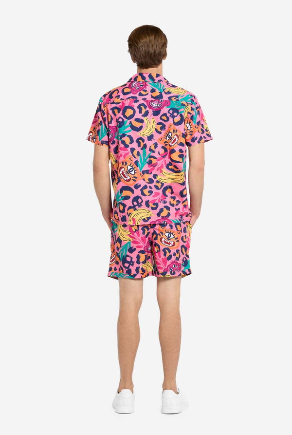Een man die een levendige tweedelige zomeroutfit draagt ​​met een opvallende tropische print. Het overhemd met korte mouwen en de bijpassende shorts hebben een levendig ontwerp met neonroze, oranje en gele luipaardprints, bananen, groene palmbladeren en eigenzinnige cartoongezichten. De outfit is nonchalant gestyled met een groen T-shirt eronder en is perfect voor zomerfeestjes, vakanties of plezier bij het zwembad. Uitzicht vanaf de achterkant.