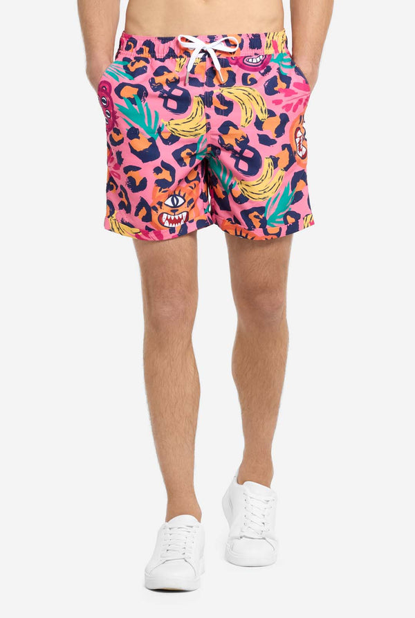 Een man die een levendige tweedelige zomeroutfit draagt ​​met een opvallende tropische print. Het overhemd met korte mouwen en de bijpassende shorts hebben een levendig ontwerp met neonroze, oranje en gele luipaardprints, bananen, groene palmbladeren en eigenzinnige cartoongezichten. De outfit is nonchalant gestyled met een groen T-shirt eronder en is perfect voor zomerfeestjes, vakanties of plezier bij het zwembad. Close-up van de korte broek.