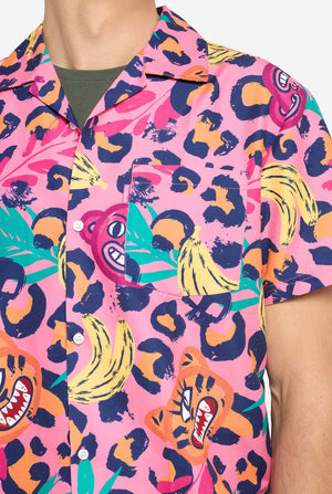 Een man die een levendige tweedelige zomeroutfit draagt ​​met een opvallende tropische print. Het overhemd met korte mouwen en de bijpassende shorts hebben een levendig ontwerp met neonroze, oranje en gele luipaardprints, bananen, groene palmbladeren en eigenzinnige cartoongezichten. De outfit is nonchalant gestyled met een groen T-shirt eronder en is perfect voor zomerfeestjes, vakanties of plezier bij het zwembad. Borst close-up.