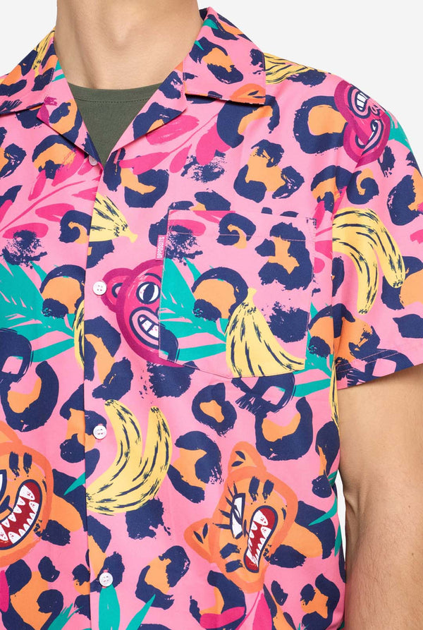 Een man die een levendige tweedelige zomeroutfit draagt ​​met een opvallende tropische print. Het overhemd met korte mouwen en de bijpassende shorts hebben een levendig ontwerp met neonroze, oranje en gele luipaardprints, bananen, groene palmbladeren en eigenzinnige cartoongezichten. De outfit is nonchalant gestyled met een groen T-shirt eronder en is perfect voor zomerfeestjes, vakanties of plezier bij het zwembad. Borst close-up.
