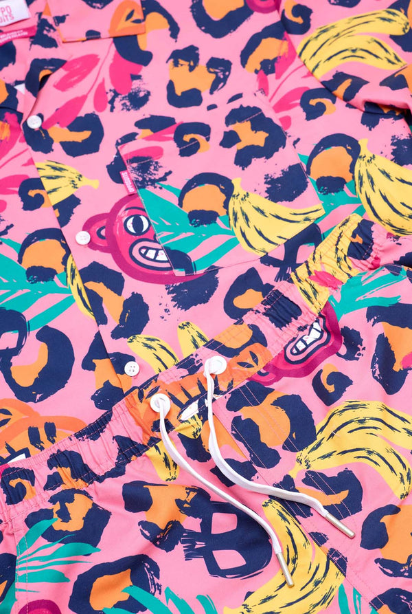 Afbeelding van een levendige tweedelige zomeroutfit met een opvallende tropische print. Het overhemd met korte mouwen en de bijpassende shorts hebben een levendig ontwerp met neonroze, oranje en gele luipaardprints, bananen, groene palmbladeren en eigenzinnige cartoongezichten. De outfit is nonchalant gestyled met een groen T-shirt eronder en is perfect voor zomerfeestjes, vakanties of plezier bij het zwembad. Close-up.