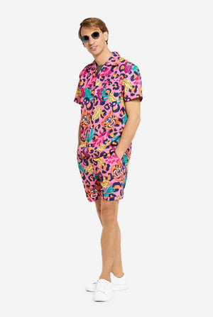 Een man in een levendige tweedelige zomeroutfit met een opvallende tropische print en een zonnebril. Het overhemd met korte mouwen en de bijpassende shorts hebben een levendig ontwerp met neonroze, oranje en gele luipaardprints, bananen, groene palmbladeren en eigenzinnige cartoon gezichten. De outfit is nonchalant gestyled met een groen T-shirt eronder en is perfect voor zomerfeestjes, vakanties of plezier bij het zwembad.