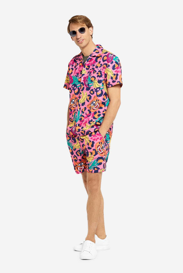 Een man in een levendige tweedelige zomeroutfit met een opvallende tropische print en een zonnebril. Het overhemd met korte mouwen en de bijpassende shorts hebben een levendig ontwerp met neonroze, oranje en gele luipaardprints, bananen, groene palmbladeren en eigenzinnige cartoon gezichten. De outfit is nonchalant gestyled met een groen T-shirt eronder en is perfect voor zomerfeestjes, vakanties of plezier bij het zwembad.