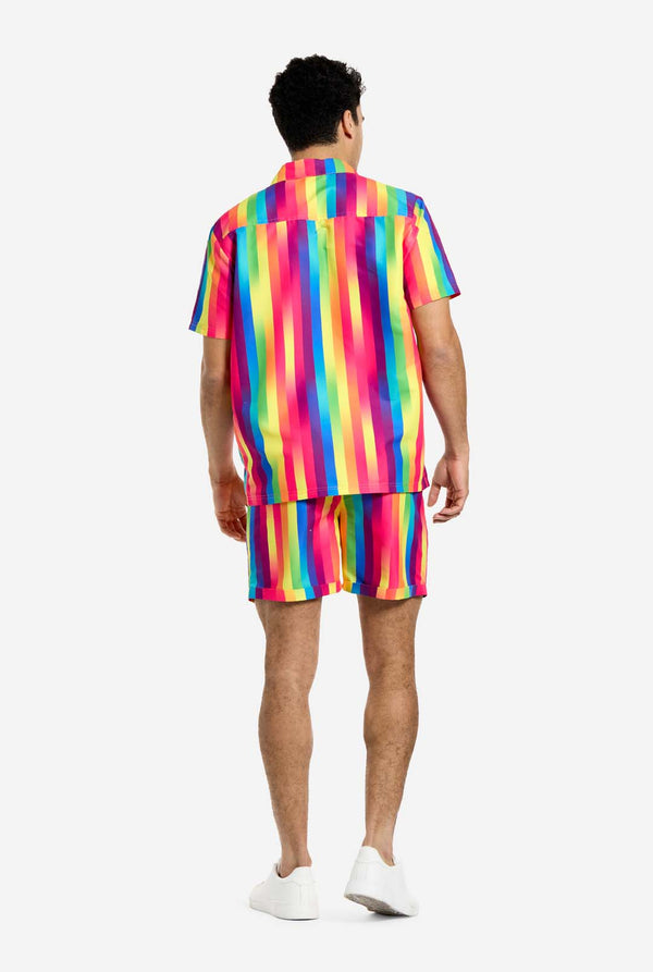 Een man draagt ​​een verticaal gestreepte regenboogkleurige zomerset, bestaande uit een overhemd en een korte broek. Hij kijkt weg van de camera en laat de achterkant van de zomerset zien.