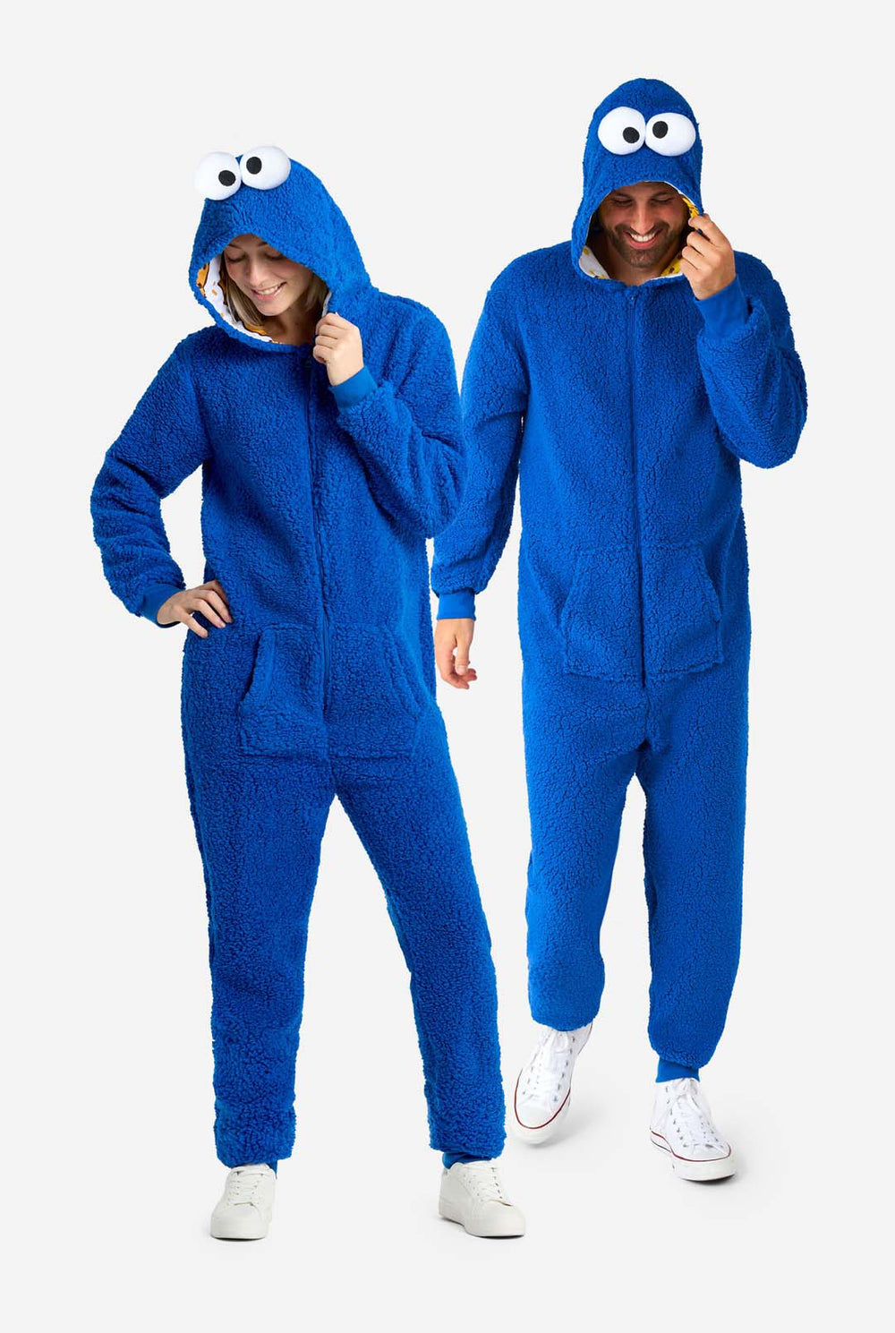 Man en vrouw dragen blauwe pluche Cookie Monster Adult onesie voor volwassenen. Staan naast elkaar, capuchon vasthoudend met de hand.