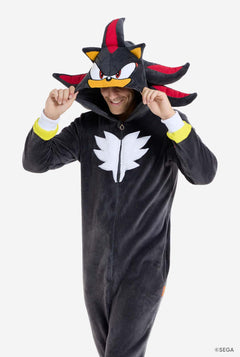 Een man draagt ​​een pluchen zwarte onesie, geïnspireerd op het populaire egelpersonage Shadow, met puntige rode en zwarte accenten op de capuchon, een wit embleem op de borst en gele details op de polsen. De onesie is gemaakt van zacht fleecemateriaal en biedt comfort en een speels ontwerp voor cosplay, loungen of thema-evenementen. De persoon glimlacht terwijl hij de capuchon aantrekt, waardoor het herkenbare gezicht en het opvallende ontwerp van het personage zichtbaar zijn.