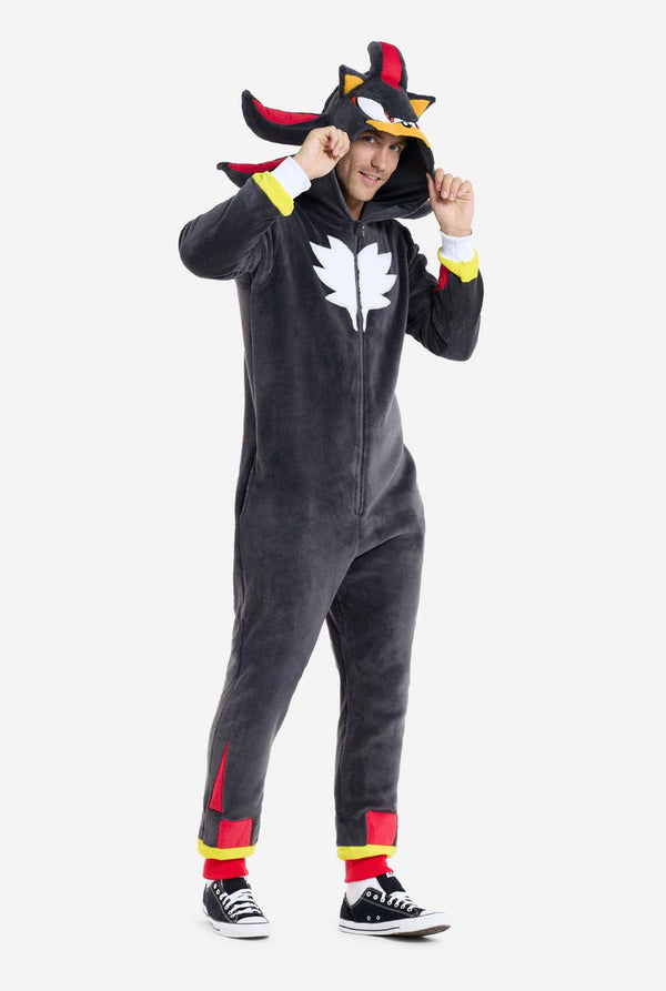 Een man met een pluchen zwarte onesie geïnspireerd op het populaire egelkarakter Shadow, met puntige rode en zwarte accenten op de capuchon, een wit embleem op de borst en gele polsdetails. De onesie is gemaakt van zacht fleecemateriaal en biedt comfort en een speels ontwerp voor cosplay, loungen of thema-evenementen. De persoon glimlacht terwijl hij de capuchon vasthoudt, wat het herkenbare gezicht en het opvallende ontwerp van het personage laat zien. Perfect voor fans van gaming of geanimeerde karakters.