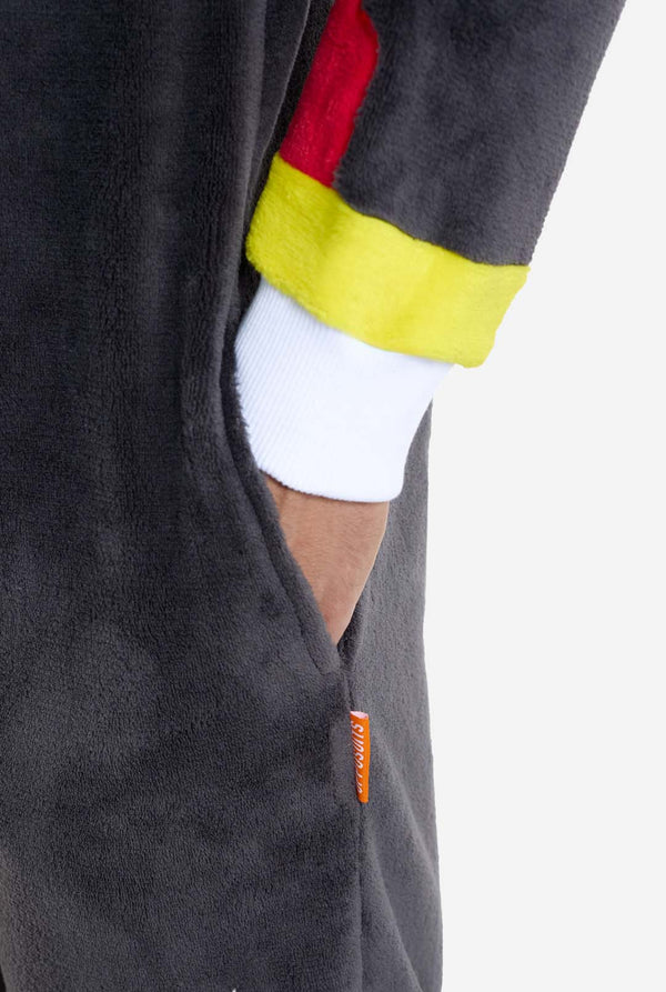 Een man met een pluchen zwarte onesie geïnspireerd op het populaire egelkarakter Shadow, met puntige rode en zwarte accenten op de capuchon, een wit embleem op de borst en gele polsdetails. De onesie is gemaakt van zacht fleecemateriaal en biedt comfort en een speels ontwerp voor cosplay, loungen of thema-evenementen.  Perfect voor fans van gaming of geanimeerde karakters. Mouw dicht met de hand in de zak.