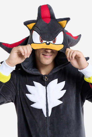 Een man met een pluchen zwarte onesie geïnspireerd op het populaire egelkarakter Shadow, met puntige rode en zwarte accenten op de capuchon, een wit embleem op de borst en gele polsdetails. De onesie is gemaakt van zacht fleecemateriaal en biedt comfort en een speels ontwerp voor cosplay, loungen of thema-evenementen. De persoon glimlacht terwijl hij de capuchon aanpast, wat het herkenbare gezicht en het opvallende ontwerp van het personage laat zien. Perfect voor fans van gaming of geanimeerde karakters. 
