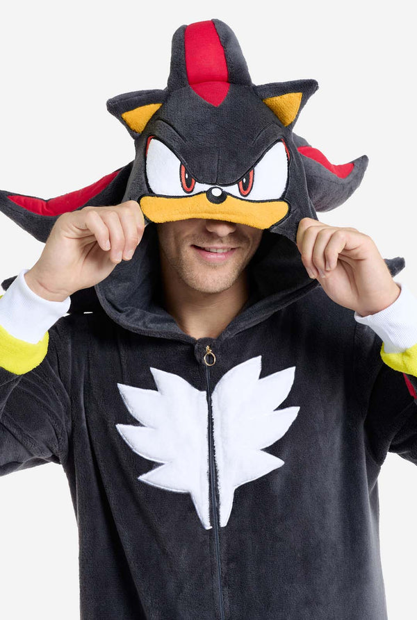 Een man met een pluchen zwarte onesie geïnspireerd op het populaire egelkarakter Shadow, met puntige rode en zwarte accenten op de capuchon, een wit embleem op de borst en gele polsdetails. De onesie is gemaakt van zacht fleecemateriaal en biedt comfort en een speels ontwerp voor cosplay, loungen of thema-evenementen. De persoon glimlacht terwijl hij de capuchon aanpast, wat het herkenbare gezicht en het opvallende ontwerp van het personage laat zien. Perfect voor fans van gaming of geanimeerde karakters. 