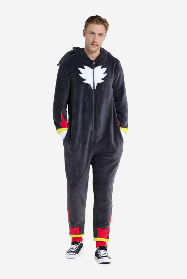 Een man met een pluchen zwarte onesie geïnspireerd op het populaire egelkarakter Shadow, met puntige rode en zwarte accenten op de capuchon, een wit embleem op de borst en gele polsdetails. De onesie is gemaakt van zacht fleecemateriaal en biedt comfort en een speels ontwerp voor cosplay, loungen of thema-evenementen. De persoon glimlacht terwijl de kap op zijn rug ligt. Perfect voor fans van gaming of geanimeerde karakters. Volledig lichaamsbeeld.