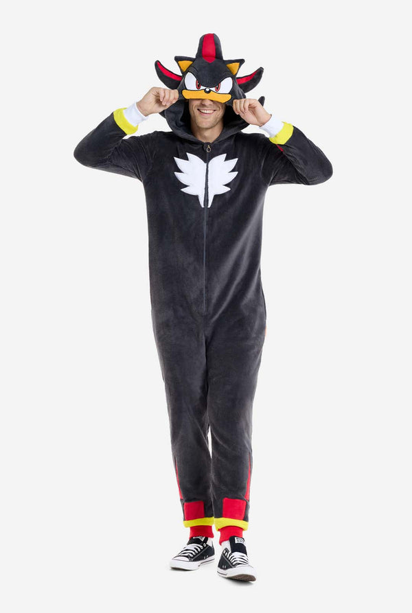 Een man met een pluchen zwarte onesie geïnspireerd op het populaire egelkarakter Shadow, met puntige rode en zwarte accenten op de capuchon, een wit embleem op de borst en gele polsdetails. De onesie is gemaakt van zacht fleecemateriaal en biedt comfort en een speels ontwerp voor cosplay, loungen of thema-evenementen. De persoon glimlacht terwijl hij de capuchon aanpast, wat het herkenbare gezicht en het opvallende ontwerp van het personage laat zien. Perfect voor fans van gaming of geanimeerde karakters. 