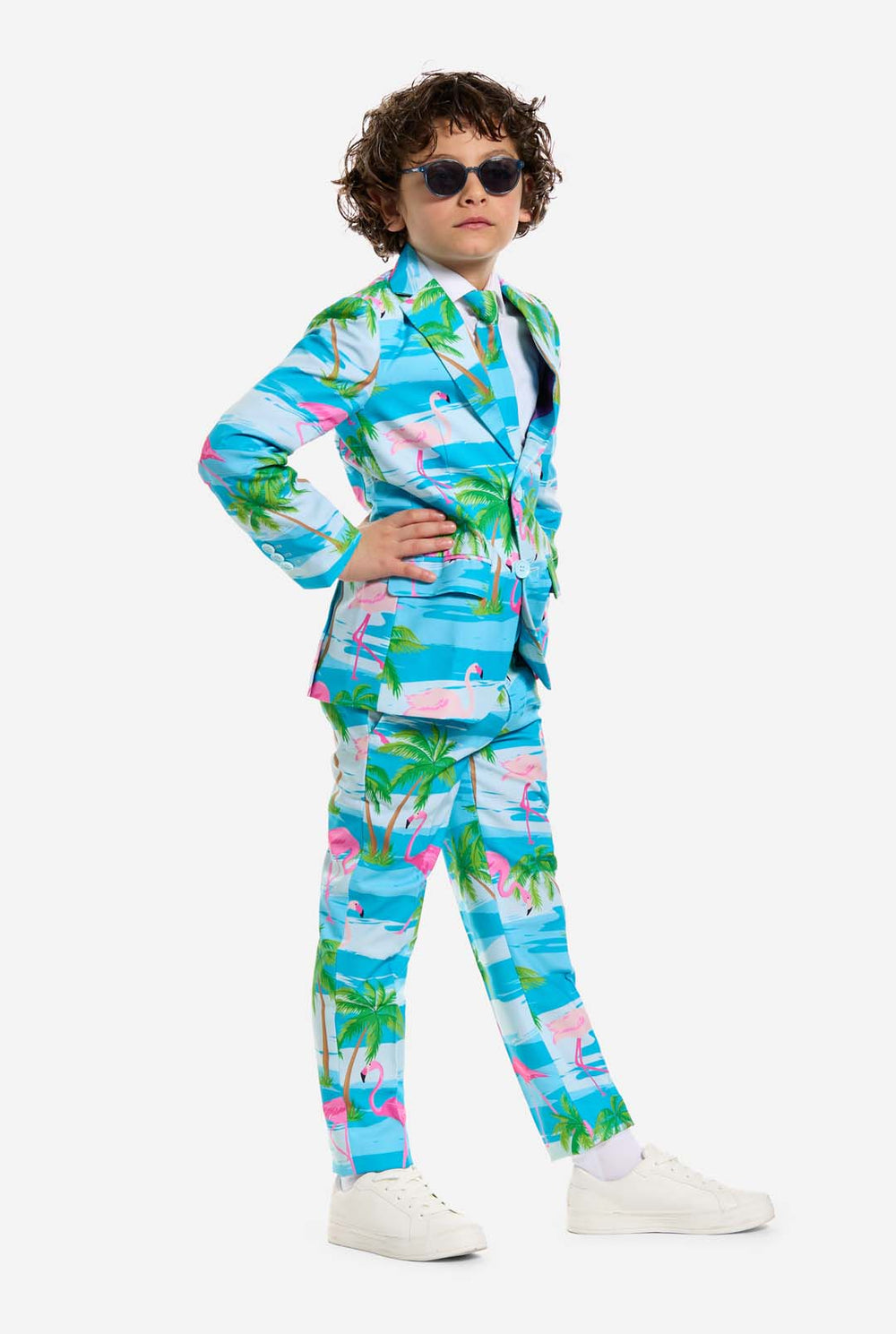 Op deze afbeelding draagt ​​een kind een lichtblauw jongenspak met een tropische print en een wit shirt. Het pak heeft een blauwe print die lijkt op water en roze flamingo's en groene palmbomen.