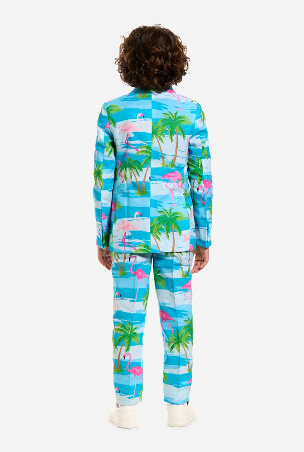 Op deze afbeelding draagt ​​een kind een lichtblauw jongenspak met een tropische print en een wit shirt. Het pak heeft een blauwe print die lijkt op water en roze flamingo's en groene palmbomen. Beeld van de achterkant.