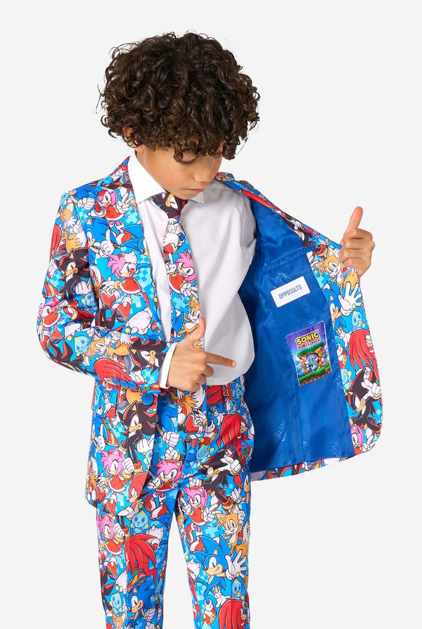 Jonge jongen draagt een levendig Sonic-tienerpak met kleurrijke opdrukken van Sonic the Hedgehog-personages zoals Sonic, Knuckles, Tails en Amy, gecombineerd met een bijpassende stropdas en wit overhemd. Houdt zijkant van het jasje open
