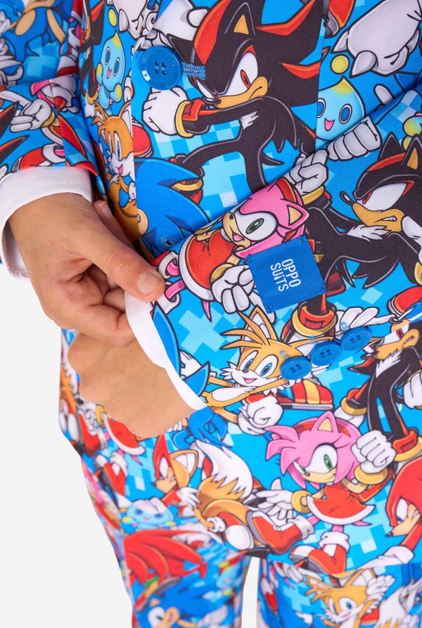 Jonge jongen draagt een levendig Sonic-tienerpak met kleurrijke opdrukken van Sonic the Hedgehog-personages zoals Sonic, Knuckles, Tails en Amy, gecombineerd met een bijpassende stropdas en wit overhemd. Gefocust op de mouwen