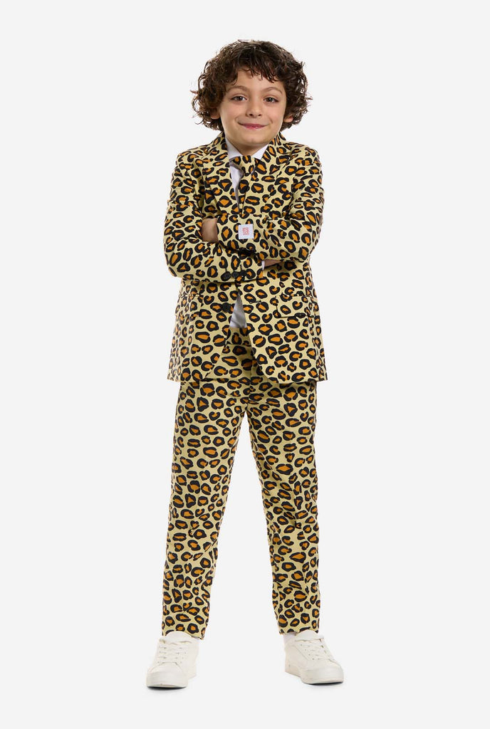 Op deze afbeelding draagt ​​een kind een jongenspak met all-over panterprint en een wit shirt. Het pak is beige met bruine en zwarte gevlekte panterprint. Pakken bestaan ​​uit een jasje, broek en clip-on stropdas. Armen over elkaar.