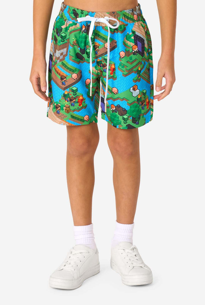 Een jonge jongen draagt een kleurrijke zomeroutfit met een Minecraft Overworld print, met pixelachtige landschappen, personages en elementen uit het iconische spel. Het shirt met korte mouwen en de bijpassende shorts benadrukken een levendig ontwerp geïnspireerd door het Minecraft-universum. Gefocust op het broekje