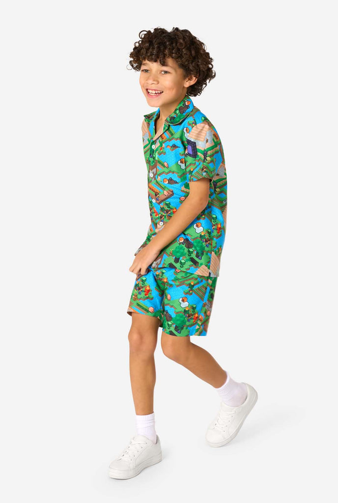 Een jonge jongen draagt een kleurrijke zomeroutfit met een Minecraft Overworld print, met pixelachtige landschappen, personages en elementen uit het iconische spel. Het shirt met korte mouwen en de bijpassende shorts benadrukken een levendig ontwerp geïnspireerd door het Minecraft-universum. Lopend