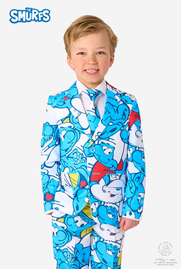 Een jongetje draagt ​​een kleurrijk Smurfenpak met daarop opvallende blauwe cartoonfiguurprints, inclusief jasje, broek en bijpassende stropdas, en lacht tegen een effen witte achtergrond.