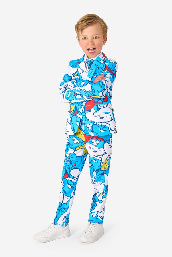 Een jongetje draagt ​​een kleurrijk Smurfenpak met daarop opvallende blauwe cartoonfiguurprints, inclusief jasje, broek en bijpassende stropdas, en lacht tegen een effen witte achtergrond. Volledig lichaam
