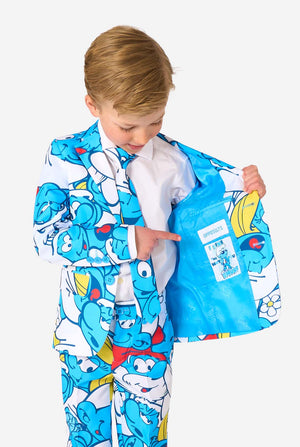 Een jongetje draagt ​​een kleurrijk Smurfenpak met daarop opvallende blauwe cartoonfiguurprints, inclusief jasje, broek en bijpassende stropdas, en lacht tegen een effen witte achtergrond. Houd een kant van het jasje open.