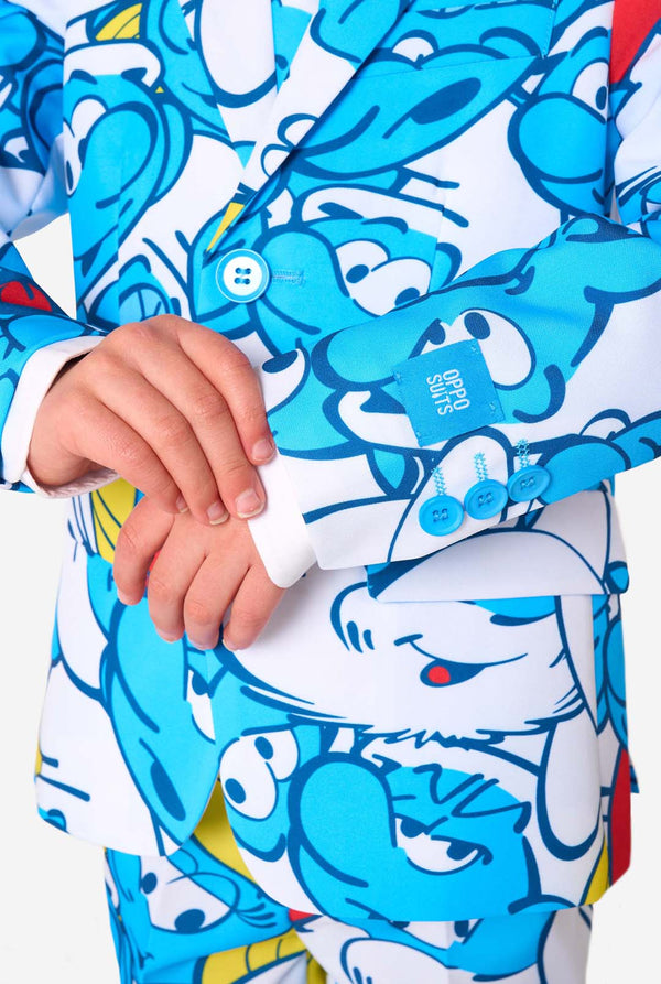 Een jongetje draagt ​​een kleurrijk Smurfenpak met daarop opvallende blauwe cartoonfiguurprints, inclusief jasje, broek en bijpassende stropdas, en lacht tegen een effen witte achtergrond. Close up van de mouwen.