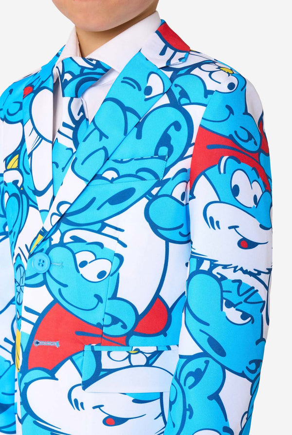 Een jongetje draagt ​​een kleurrijk Smurfenpak met daarop opvallende blauwe cartoonfiguurprints, inclusief jasje, broek en bijpassende stropdas, en lacht tegen een effen witte achtergrond. Close up van de borst.