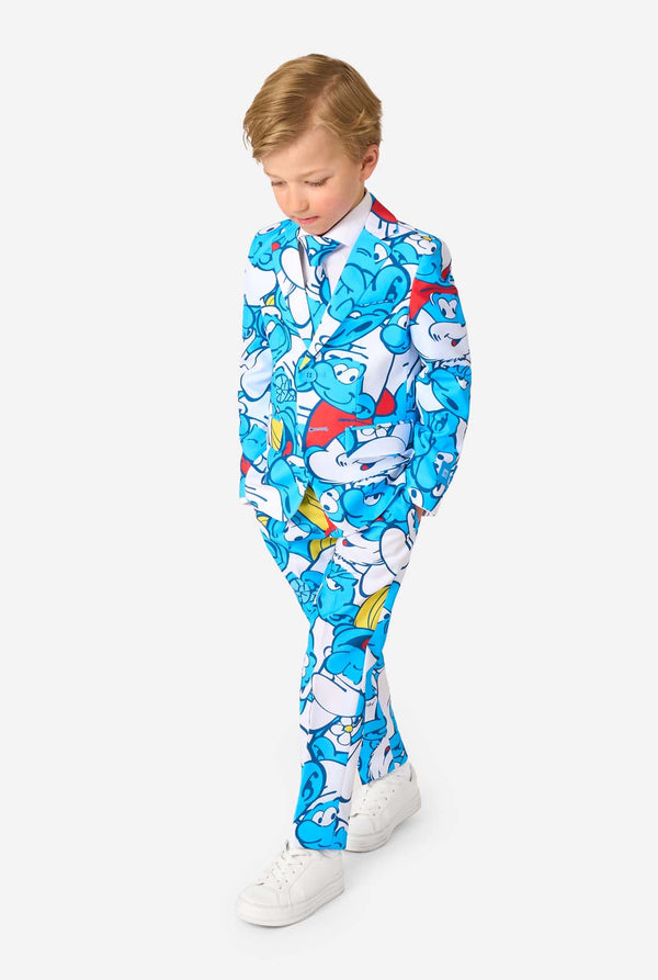 Een jongetje draagt ​​een kleurrijk Smurfenpak met daarop opvallende blauwe cartoonfiguurprints, inclusief jasje, broek en bijpassende stropdas, en lacht tegen een effen witte achtergrond. Kijkt naar de grond.