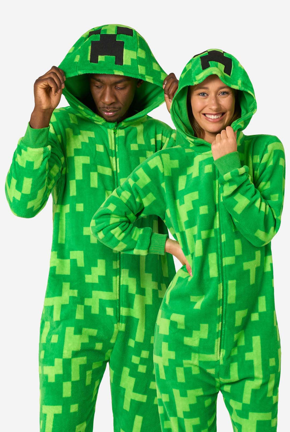 Twee mensen die groene, gepixelde onesies dragen die zijn ontworpen om op het Minecraft Creeper-personage te lijken. De onesies hebben capuchons met het iconische zwarte pixelgezicht van de Creeper. De man houdt zijn capuchon vast en kijkt naar beneden, terwijl de vrouw met één hand op de heup naar de camera lacht