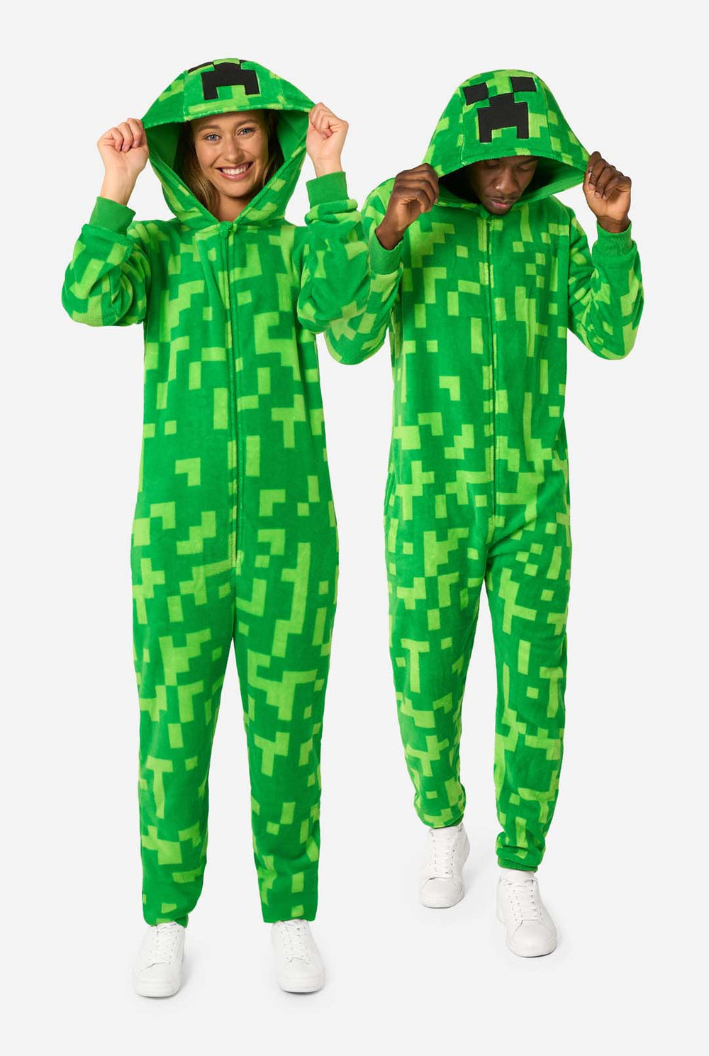 Twee mensen die groene, gepixelde onesies dragen die zijn ontworpen om op het Minecraft Creeper-personage te lijken. De onesies hebben capuchons met het iconische zwarte pixelgezicht van de Creeper. De man houdt zijn capuchon vast en kijkt naar beneden, terwijl de vrouw met één hand op de heup naar de camera lacht