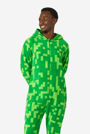 Een man die een groene, pixelachtige onesie draagt, ontworpen om op het Minecraft Creeper-personage te lijken. De onesie heeft een capuchon met het iconische zwarte pixelgezicht van de Creeper. De man heeft de capuchon op zijn rug en kijkt naar de zijkant.