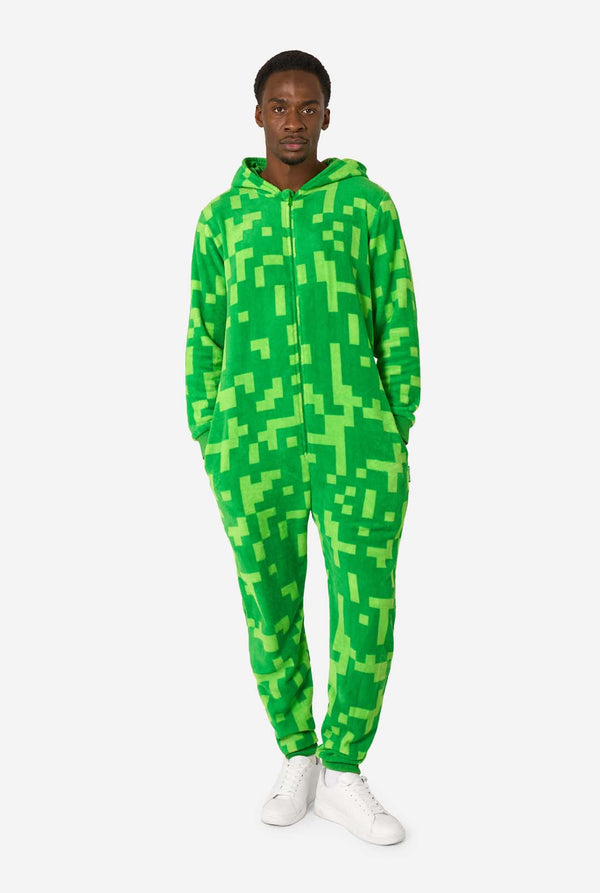 Een man die een groene, pixelachtige onesie draagt, ontworpen om op het Minecraft Creeper-personage te lijken. De onesie heeft een capuchon met het iconische zwarte pixelgezicht van de Creeper. De man heeft de kap op zijn rug en kijkt in de camera, volledig lichaamsbeeld.