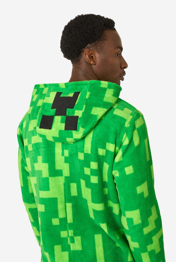 Een man die een groene, pixelachtige onesie draagt, ontworpen om op het Minecraft Creeper-personage te lijken. De onesie heeft een capuchon met het iconische zwarte pixelgezicht van de Creeper. De man kijkt opzij. Uitzicht vanaf de achterkant.