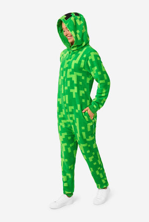 Een vrouw die een groene, pixelachtige onesie draagt, ontworpen om op het Minecraft Creeper-personage te lijken. De onesie heeft een capuchon met het iconische zwarte pixelgezicht van de Creeper. De vrouw draagt ​​de kap en kijkt naar de grond.