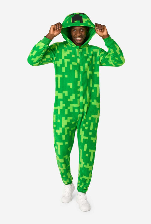 Een man die een groene, pixelachtige onesie draagt, ontworpen om op het Minecraft Creeper-personage te lijken. De onesie heeft een capuchon met het iconische zwarte pixelgezicht van de Creeper. De man draagt ​​de kap en kijkt in de camera