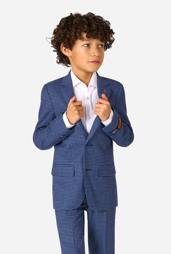 Een jongen, gekleed in een marineblauw en blauw geruit pak met een wit overhemd. Het pak heeft een moderne pasvorm met twee knopen en klepzakken. Hij staat tegen een effen witte achtergrond.