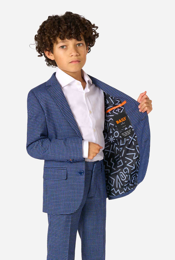 Een jongen, gekleed in een marineblauw en blauw geruit pak met een wit overhemd. Het pak heeft een moderne pasvorm met twee knopen en klepzakken. Hij staat tegen een effen witte achtergrond. Houdt een kant van het jasje open.