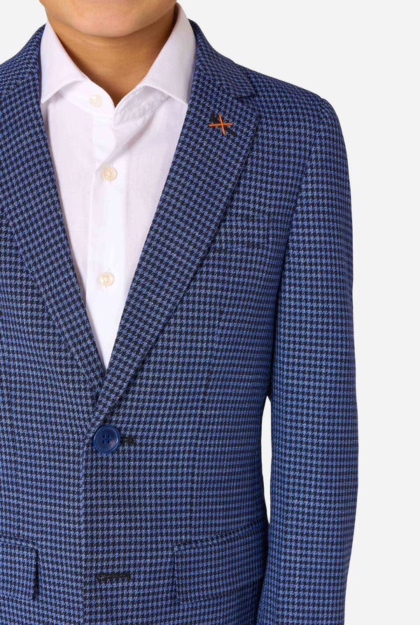 Een jongen, gekleed in een marineblauw en blauw geruit pak met een wit overhemd. Het pak heeft een moderne pasvorm met twee knopen en klepzakken. Hij staat tegen een effen witte achtergrond. Close-up van de borst