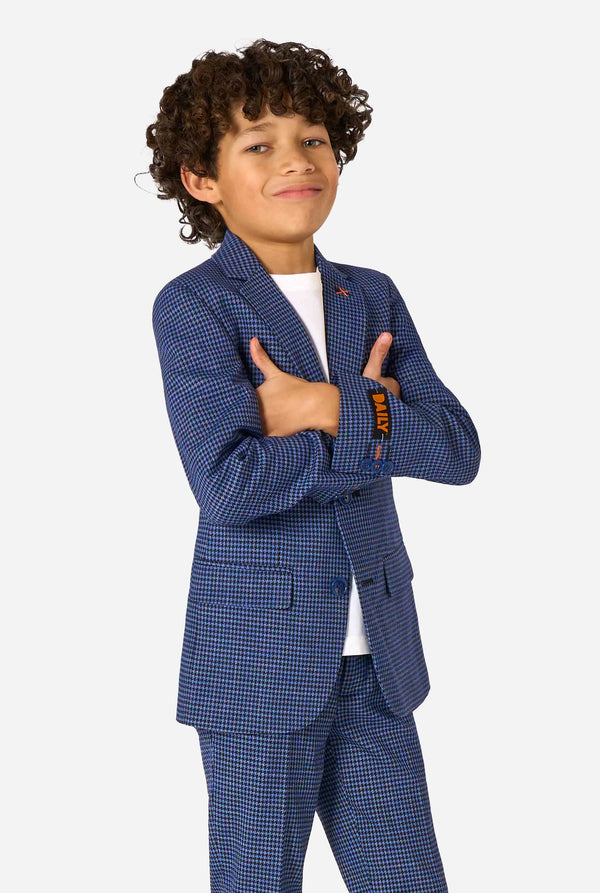 Een jongen, gekleed in een marineblauw en blauw geruit pak met een wit overhemd. Het pak heeft een moderne pasvorm met twee knopen en klepzakken. Hij staat tegen een effen witte achtergrond.